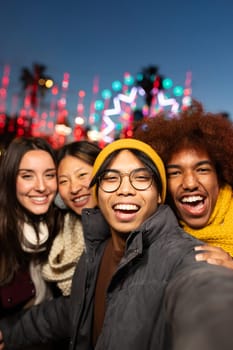 Multiracial young friends laughing and having fun together taking vertical selfie during winter market at night looking at camera. Friendship concept.