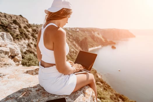 Digital nomad, Business woman working on laptop by the sea. Pretty lady typing on computer by the sea at sunset, makes a business transaction online from a distance. Freelance remote work on vacation