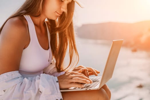 Digital nomad, Business woman working on laptop by the sea. Pretty lady typing on computer by the sea at sunset, makes a business transaction online from a distance. Freelance remote work on vacation