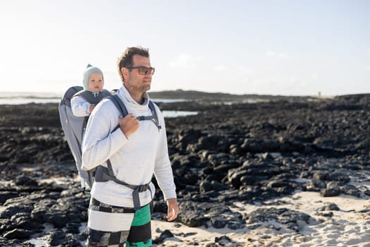 Young father carrying his infant baby boy son in backpack on black rock volcanic beach on Lanzarote island, Spain. Family travel and winter vacation concept