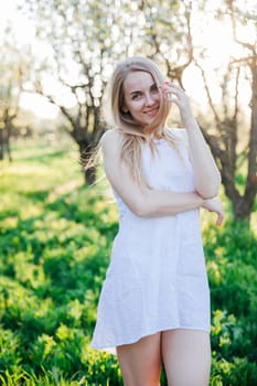 a blonde woman in white dress walks in park flowering trees garden