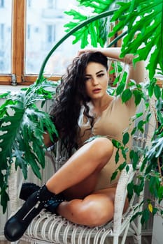 a brunette woman in a beige bodysuit sits on a chair near green plants