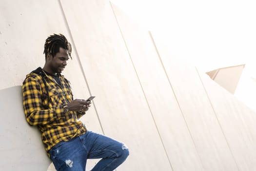 smiling young black man writing a message on his phone leaning on a modern concrete structure, concept of lifestyle and technology
