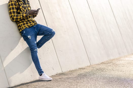 unrecognizable young black man writing a message on his phone leaning on a modern concrete structure, concept of lifestyle and technology