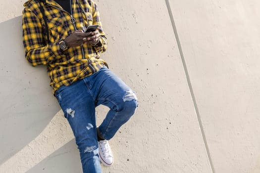 unrecognizable young african man writing a message on his phone leaning on a modern concrete structure, concept of lifestyle and technology