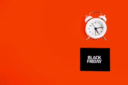 white alarm clock and an inscription on a black square - black Friday on a red background. The topic of sales. Flat lay.