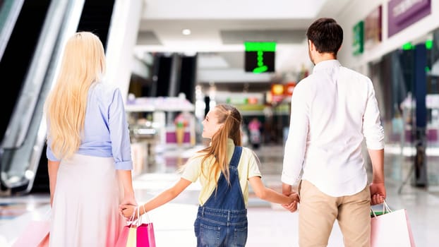 A family of three walks through a shopping mall. They are holding hands, and the parents each carry a shopping bag. The young girl is looking back at something behind them