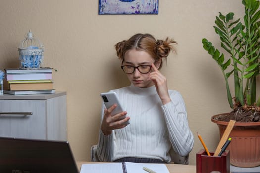 beautiful girl in glasses looks thoughtfully at the smartphone screen while sitting at her desk. soft focus