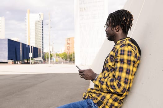 young african man with his phone leaning on a modern concrete structure at the city, concept of lifestyle and technology