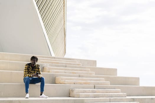 young black african-american man sitting on a concrete staircase with his phone, concept of lifestyle and technology, copy space for text