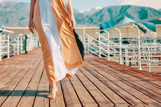 Legs of a woman in a dress walking along the pier on the beach by the sea in a hotel while relaxing against the backdrop of high mountains