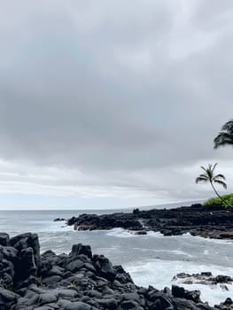 Black lava rocks stand starkly against a stormy gray sky as waves crash against the shore, creating a scene of natural beauty and tranquility