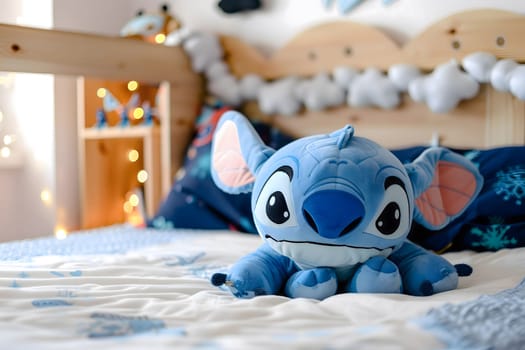 A blue Stitch plush toy sits on a bed with white bedding. The toy has big eyes, a smile and a fluffy texture. The bed is decorated with white cloud and star decorations.