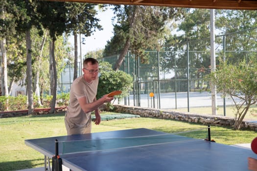 young guy plays ping pong outdoors, on table tennis table, actively hit a yellow ball with an orange racket, an unforgettable summer vacation in Turkey