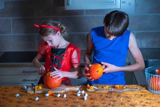 Little girl in a Halloween devil costume and her brother cut out eyes on a pumpkin with a sharp knife in a darkened room, a traditional autumn holiday.