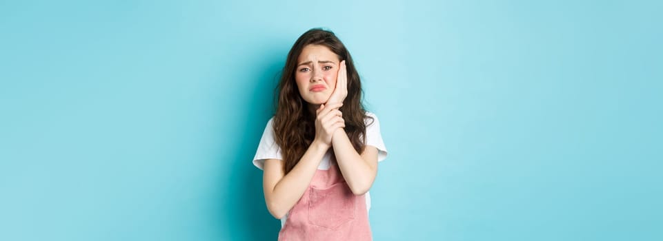 Image of frowning upset girl touching swallen cheek, having toothache, complaining on tooth decay, need to see dentist, standing against blue background.