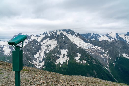 A drone flies over a mountain valley in the Caucasus Mountains. The valley is filled with green trees and a winding river. In the distance, a snow-capped peak rises above the valley.