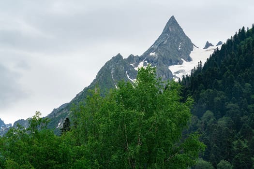 A drone flies over a mountain valley in the Caucasus Mountains. The valley is filled with green trees and a winding river. In the distance, a snow-capped peak rises above the valley.