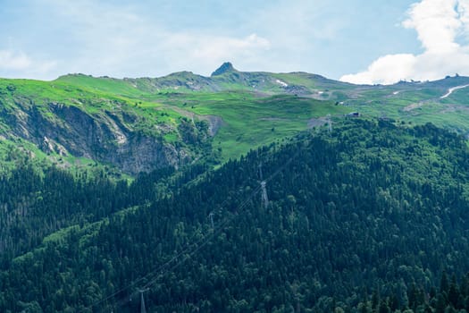 A drone flies over a mountain valley in the Caucasus Mountains. The valley is filled with green trees and a winding river. In the distance, a snow-capped peak rises above the valley.