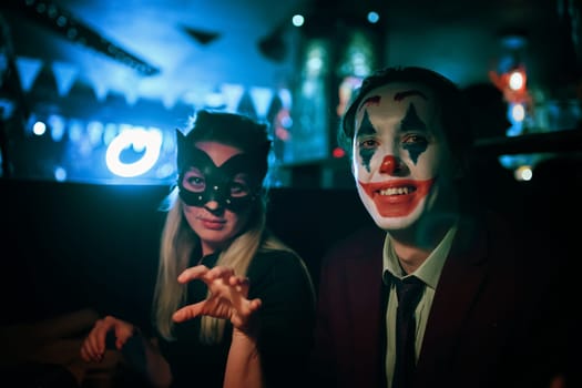 Halloween party at a nightclub. Woman in cat mask and guy with joker makeup sits at table in pub. All Saints' Night. Garland lights on the background. Photo card with copy space.