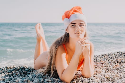 A young woman wearing a red swimsuit and a Santa hat lies on a beach with small pebbles. She gazes at the camera with a relaxed and happy expression.