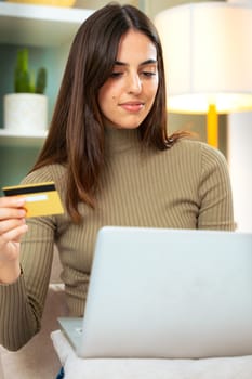 Pretty young caucasian woman shopping online with credit card and laptop while sitting on the couch at home.Vertical