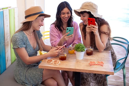 Group of smiling multiethnic women enjoying vacation in a restaurant. Beautiful and cheerful Generation Z girls browsing the internet with smartphones