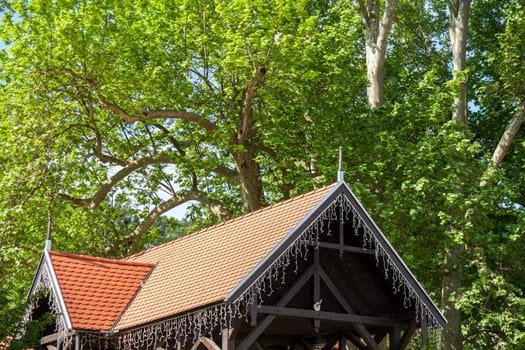 Rustic wooden structure with a terracotta-tiled roof, adorned with string lights, surrounded by lush green trees.
