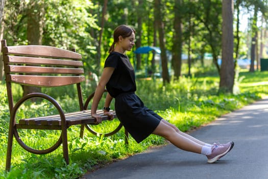 Beautiful young sporty woman in black t-shirt, black shorts and pink trainers warming up exercising triceps dip doing push-ups from bench among trees before running.