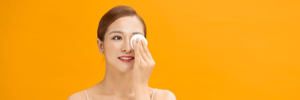 Beautiful young woman cleaning her face with cotton on yellow bachground, banner