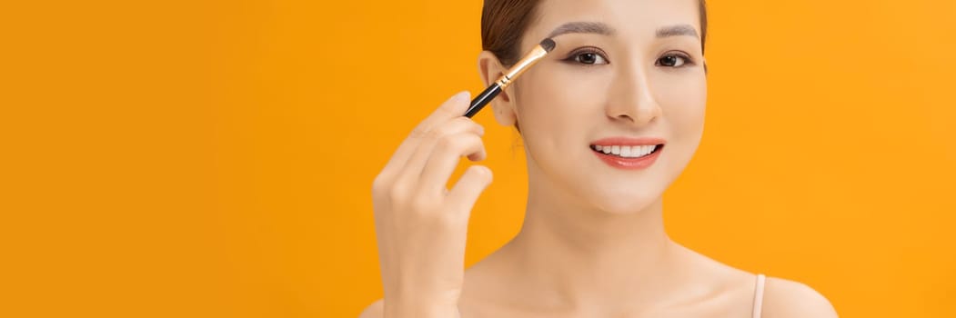 Yellow banner studio photo of a young woman using make up brush and eye shadows.