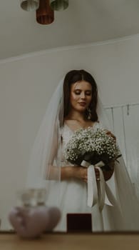 Portrait of one beautiful young Caucasian happy brunette bride with a white bouquet of flowers and boutonnieres looking thoughtfully down, reflected in the mirror, side view, close-up.
