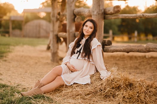 a pregnant woman in a dress in the countryside is sitting on the hay near the horses.