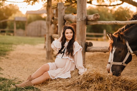 a pregnant woman in a dress in the countryside is sitting on the hay near the horses.