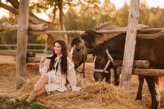 a pregnant woman in a dress in the countryside is sitting on the hay near the horses.