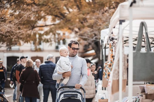 Father walking carrying his infant baby boy child and pushing stroller in crowd of people wisiting sunday flea market in Malaga, Spain