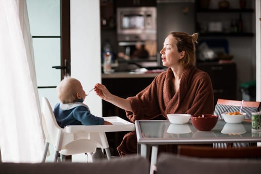 Mother wearing bathrope spoon feeding her infant baby boy child sitting in high chair at the dining table in kitchen at home in the morning.