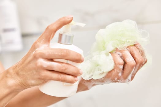 female hands pouring liquid moisturizing soap shower gel from white cosmetic bottle on bath puff in bathroom