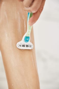 Young woman shaving her legs with razor in white shower, close-up