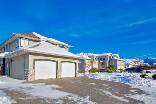 Double garage of big luxury house with driveway and front yard in snow. Street of residential houses on winter sunny day