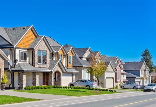 A perfect neighborhood. A street of family houses in suburban area of Vancouver with concrete side walk and asphalt road in front. Residential houses with cars parked on driveways.