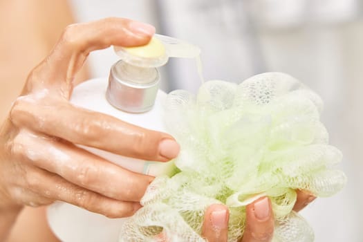 female hands pouring liquid moisturizing soap shower gel from white cosmetic bottle on sponge in bathroom