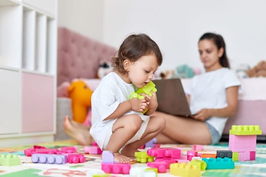 cute little baby girl playing with toys blocks and mother mother working on laptop from home