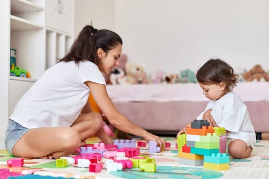 cute baby girl and her loving mother playing with toy bricks in the children room, early development