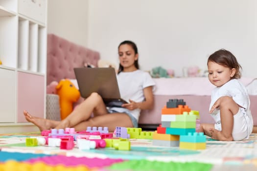 cute little baby girl playing with toys blocks and mother working on laptop from home
