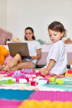cute little baby girl playing with toys blocks and mom sitting on floor with laptop in room