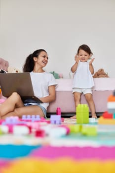 cute little baby girl playing with toys blocks and mother sitting on floor with laptop in room