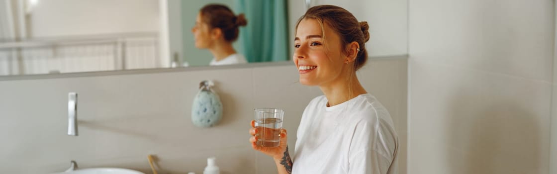 Smiling young woman drinking glass of water in the bathroom. Morning beauty routine