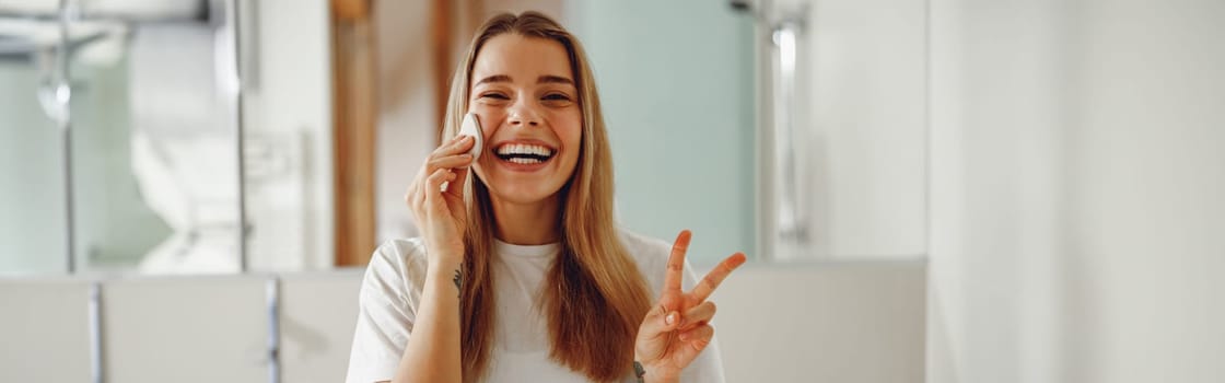 Woman wiping her face with cotton pad in the bathroom and showing sign peace. Skin care concept