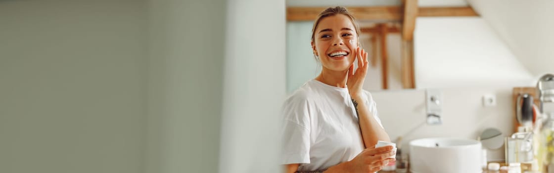 Beautiful young woman taking care of skin by applying moisturizer cream in bathroom